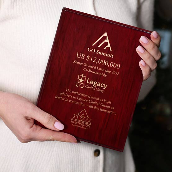 A person holding a rosewood plaque with engraved beige text and logos, including GO Summit, Legacy Capital Group, and Huntington Financial & Asset.