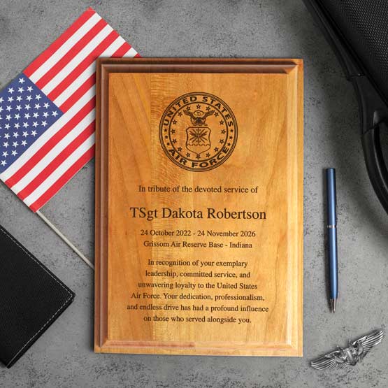 A custom solid cherry plaque, featuring the United States Air Force seal at the top and a memorial text for TSgt Dakota Robertson. The plaque is photographed on a gray textured surface with a small American flag, a pen, and a wing emblem nearby.
