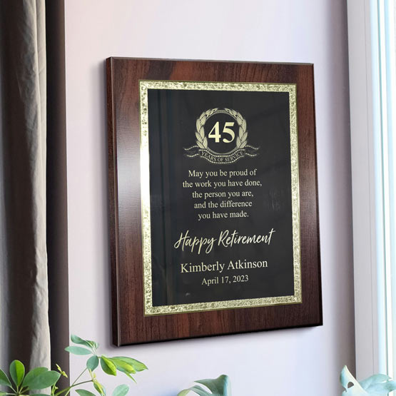 A personalized 45th retirement milestone plaque with a cherry wood board and a black metal plate with decorative gold boarder. The plaque features a gold laurel wreath and ’45 Years Service’ emblem, displayed on a white wall with a green plant in the corner. 