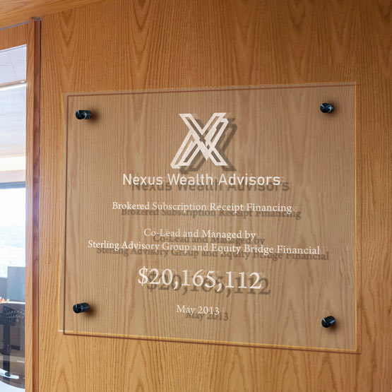 A rectangular clear acrylic sign mounted on a light wood door, featuring engraved text for Nexus Wealth Advisors and a large dollar amount, secured with four black standoffs.