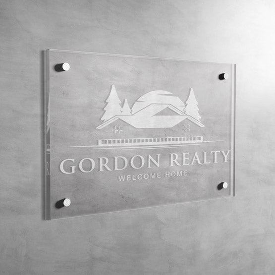 Clear Rectangle Acrylic Sign With Frosted Engravings Of Realty Company Logo And Name And Slogan Mounted Onto Gray Office Wall