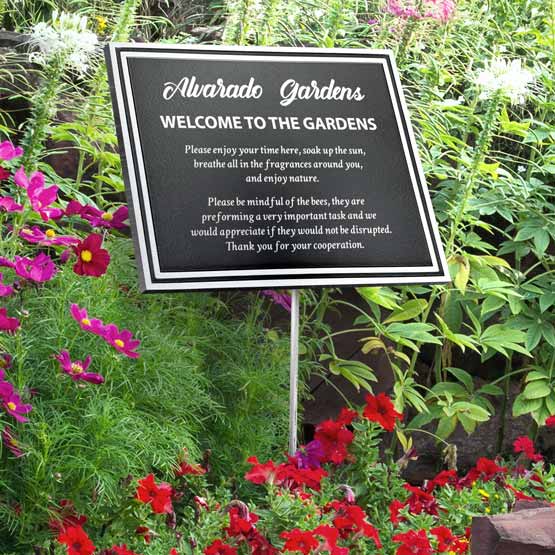 A black cast aluminum sign with a white boarder and white engraved text reading ‘Alvarado Gardens Welcome to the Gardens’ displayed in a garden bed surrounded by green foliage and red and pink flowers.