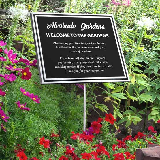 A black cast aluminum sign with a white boarder and white engraved text reading ‘Alvarado Gardens Welcome to the Gardens’ displayed in a garden bed surrounded by green foliage and red and pink flowers.