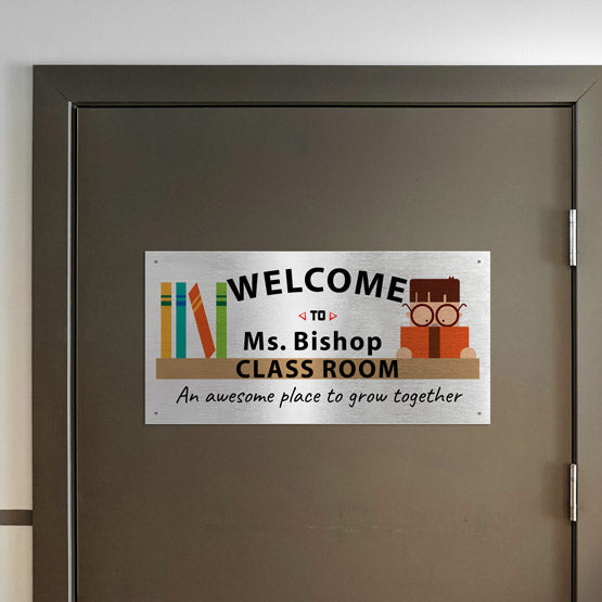 A silver metal sign with full color printing on a door. The sign is a welcome sign to Ms Bishop’s classroom attached to the door by four screws.