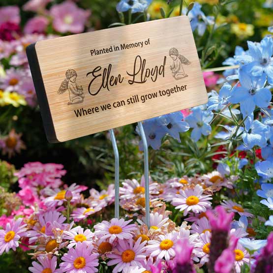 A personalized bamboo garden sign mounted on a metal stake engraved with a memorial for Ellen Lloyd, displayed in a colorful flower bed. 