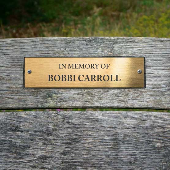 A rectangular brass metal memorial sign mounted on a weathered gray wooden bench, with black engraved text of ‘In Memory of BOBBI CARROLL’.