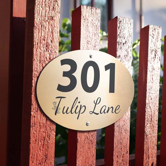 An oval bronze metal address sign mounted on a reddish-brown wooden fence, with the engraved text ‘301 Tulip Lane’.