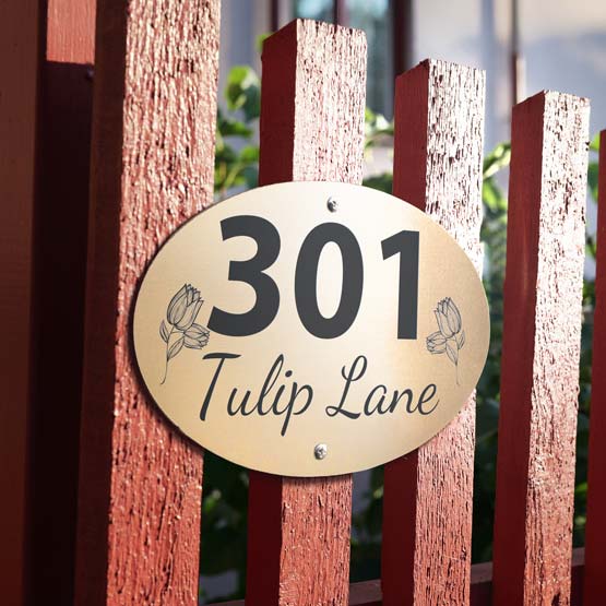 An oval bronze metal address sign mounted on a reddish-brown wooden fence, with the engraved text ‘301 Tulip Lane’.