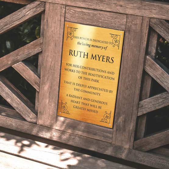 A rectangular bronze memorial sign with black engraved text dedicated to the memory of ‘RUTH MYERS’, mounted to an outdoor wooden bench.