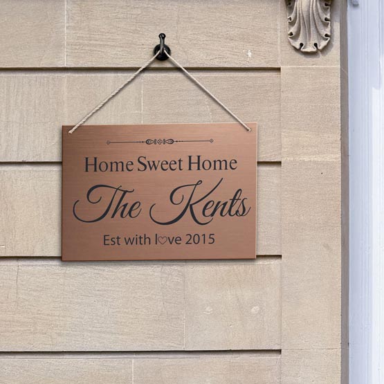 A personalized copper hanging sign with the text ‘Home Sweet Home The Kents Eat with love 2015’ with black engraved text, hanging by twine rope on the side of a house with a light green background.