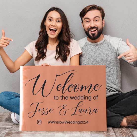 Happy Couple Sitting On Living Room Floor Holding And Pointing To Copper Sign Laser Fused With Wedding Welcome Message