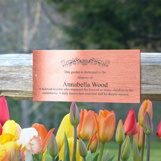 A rectangular copper memorial sign with engraved black text reading about a garden dedicated to Annabella Wood, mounted outside to a wooden fence, surrounded by colorful tulips.