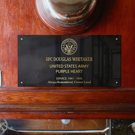 A laser metal plaque mounted on a polished wooden surface. The plaque reads 'SPC DOUGLAS WHITAKER', 'UNITED STATES ARMY', 'PURPLE HEART', and 'SERVICE: 1941 - 1945 ', with the United States Army seal above the text.