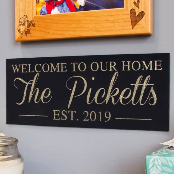 Black And Gold Faux Leather Sign Engraved With Welcome To Our Home And Family Name And Date On Wall Beneath Picture Frame