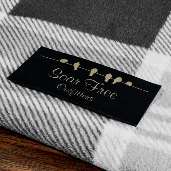 Black And Gold Faux Leather Label Engraved With Birds Art And Soar Free Outfitters Logo Fastened To Carpet On Wood Floor