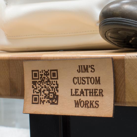 Rectangle Leather QR Tag Engraved With QR Code And Jim’s Custom Leather Works Text On Desk Edge Beneath Shoe And Purse