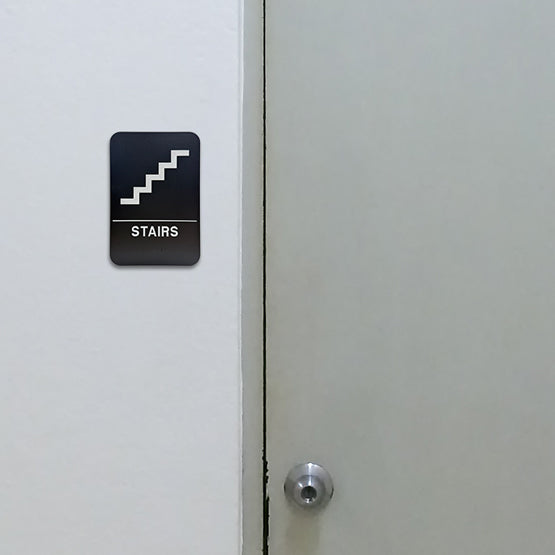 Black And White Plastic Sign With Stairs Icon Above Corresponding Text And Braille Mounted On Wall Beside Stair Access Door