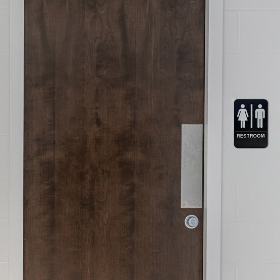 Black And White Plastic Sign With Icons Of Man And Woman For Unisex Restrooms On Wall Beside Wooden Public Restroom Door