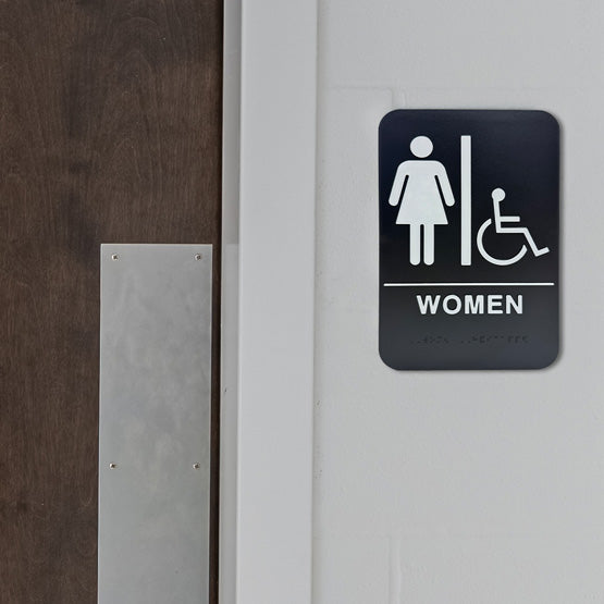 Black And White Plastic Sign With Woman And Wheelchair Icons Above Woman Text With Braille Mounted On Wall Beside Wood Door