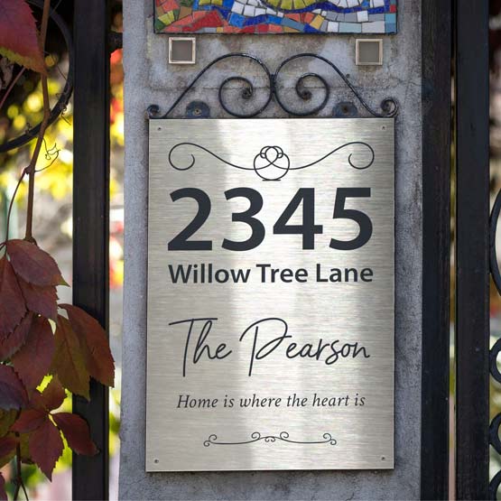 A stainless steel metal personalized house sign mounted on a stone pillar, featuring the address ‘2345 Willow Tree Land’, the name ‘The Pearson’, and the phrase ‘Home is where the heart is’ in engraved text. The sign is shown outdoors next to a black metal gate with autumn leaves hanging on the left.