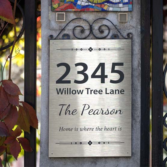 A stainless steel metal personalized house sign mounted on a stone pillar, featuring the address ‘2345 Willow Tree Land’, the name ‘The Pearson’, and the phrase ‘Home is where the heart is’ in engraved text. The sign is shown outdoors next to a black metal gate with autumn leaves hanging on the left.