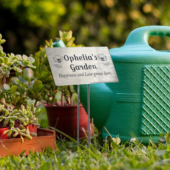A personalized stainless steel garden stake sign reading ‘Ophelia’s Garden’ with the phrase ‘Happiness and Love grows here.’ Displayed in a real garden setting next to a teal watering can.