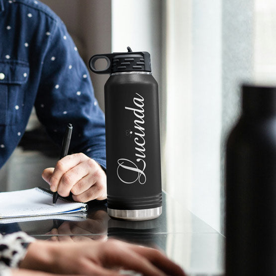 A matte black insulated water bottle with a silver stainless steel base and a script name ‘Lucinda’ engraved on the front, sitting on a reflective table next to a person writing in a notebook.