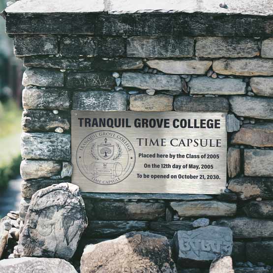 A stainless steel time capsule sign for Tranquil Grove College mounted on a stone wall, with text detailing when the capsule was placed on 2005 and when the capsule is to be opened October 21, 2030.