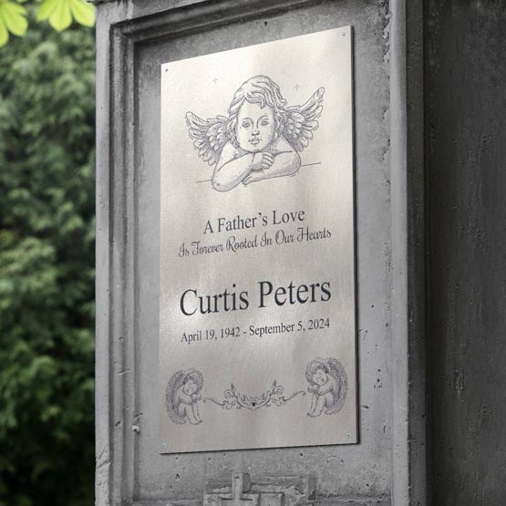 Personalized titanium memorial sign mounted to a dark stone gravestone, featuring a cherub artwork and black engraved text ‘A Father’s Love’ and ‘Curtis Peters’. Photographed outdoors with blurred green foliage in the background.