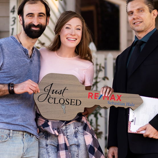 Realtor Posing Outdoors With Happy Couple Holding House Key Shaped Walnut Wood Sign Printed With Just Closed Message And Logo