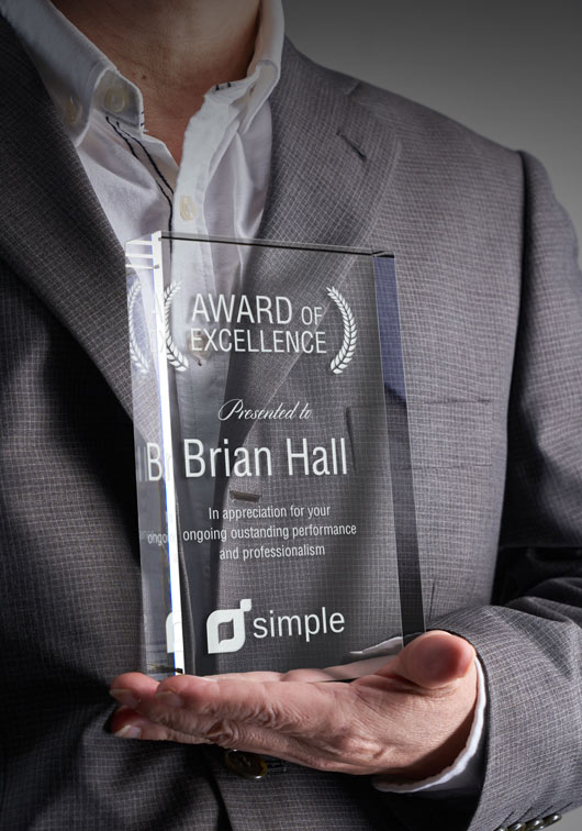 Person holding a custom standing glass award titled 'Award of Excellence' for Brian Hall.