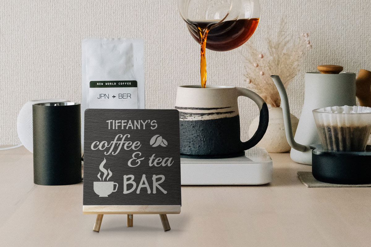 A slate tile with a customized design engraved on the front, sitting on a small wooden easel, next to a coffee bar.