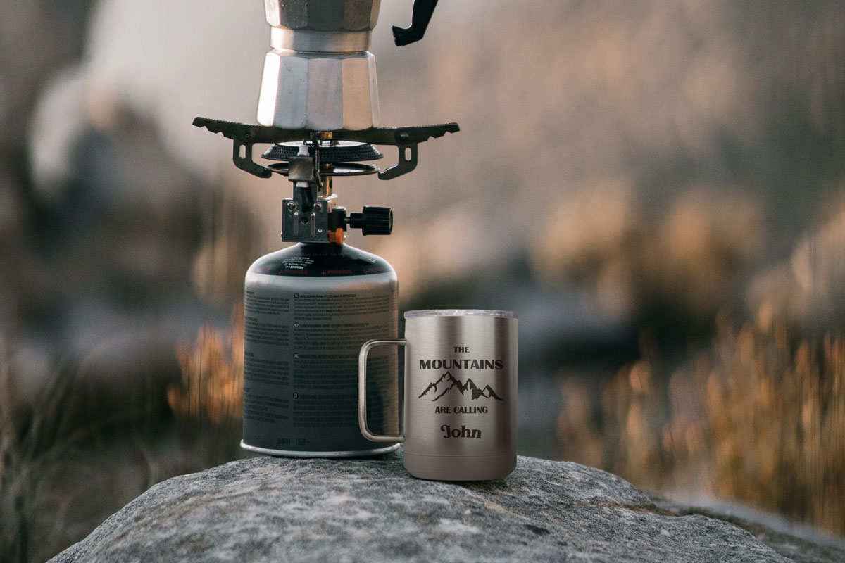 A customized silver 15oz coffee tumbler, sitting on a rock next to portable coffee machine with mountains in the background.
