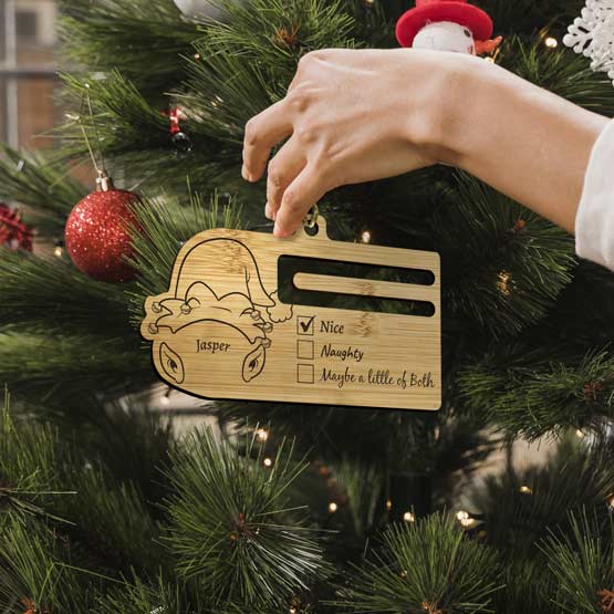 Woman’s Hand Putting Custom Bamboo Money Holder Christmas Ornament With Elf Art And Nice Checkmark Onto Tree Via Gold String