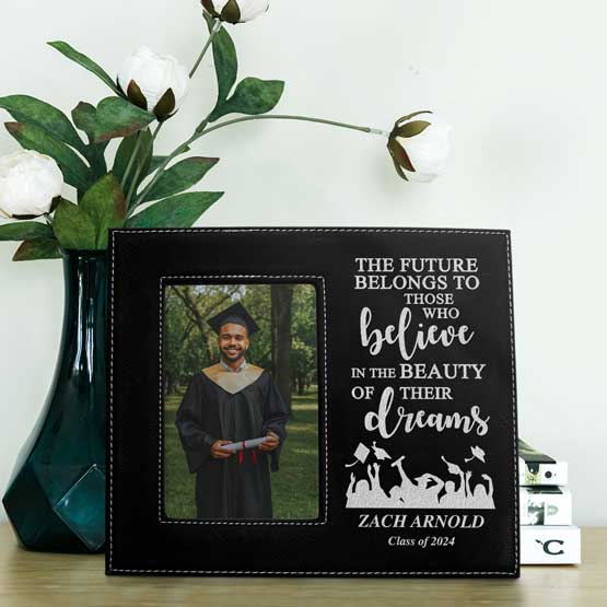 Custom Engraved Black Faux Leather Graduation Frame with Personalized Name and Class Year. Frame is on Desk near Plants and Books.