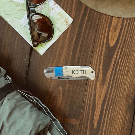 Stainless Steel Folding Knife With Blue And Bone Handle Featuring Custom Name Sitting On Wood Table With Sunglasses And Map