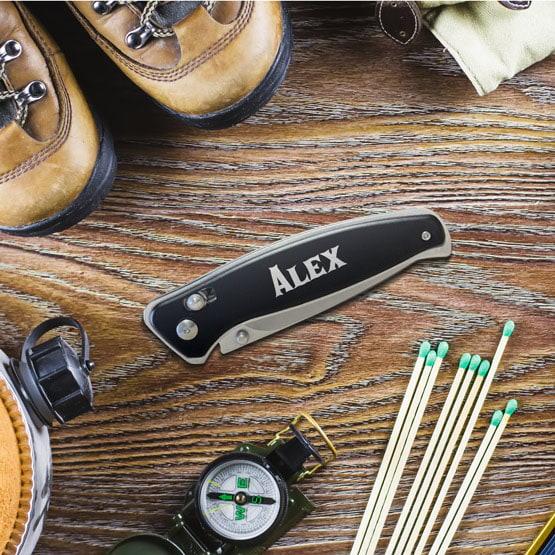 Black Folding Knife With Button Lock And Clip And Custom Name On Handle Sitting On Wood Table With Travel Essentials
