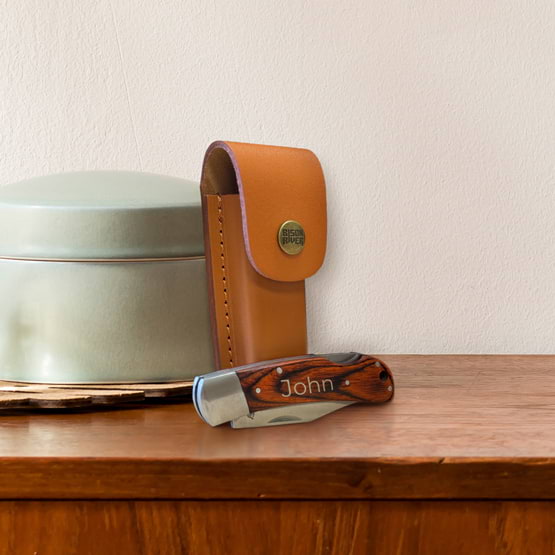 Custom Pakkawood Folding Knife Featuring Custom Name Beside Matching Leather Sheath And Sitting On Wood Dresser