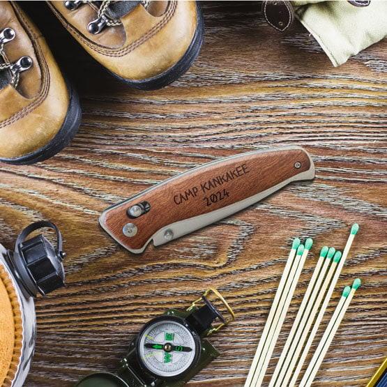 Wood Folding Knife With Button Lock And Clip Featuring Custom Camp Name And Year Sitting On Wood Table With Travel Tools