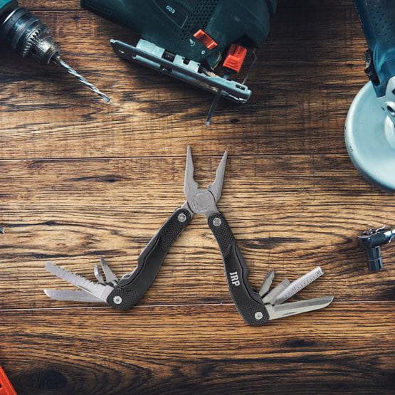 Black Multi Tool Featuring Custom Acronym With Tools Opened While Sitting On Wood Work Table With Other Tools