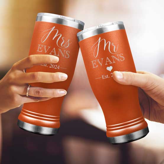 Two Custom Engraved Orange Beer Pilsner Tumblers held by woman and man with sample engraving for Mr & Mrs Evans