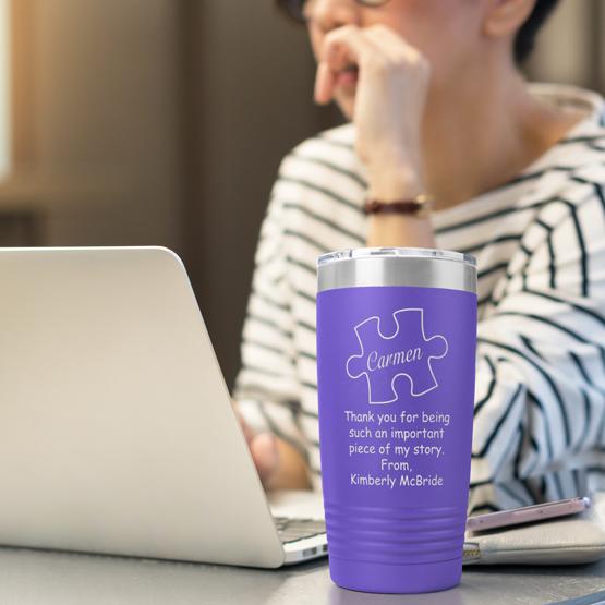 Piece of My Story 20 oz purple tumbler sitting on a table next to a woman working on a laptop. Tumbler engraved with her name