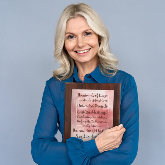 Older Blonde Woman In Blue Blouse Holding Retirement Plaque With Brown Board And Pink Watercolor Aluminum Plate