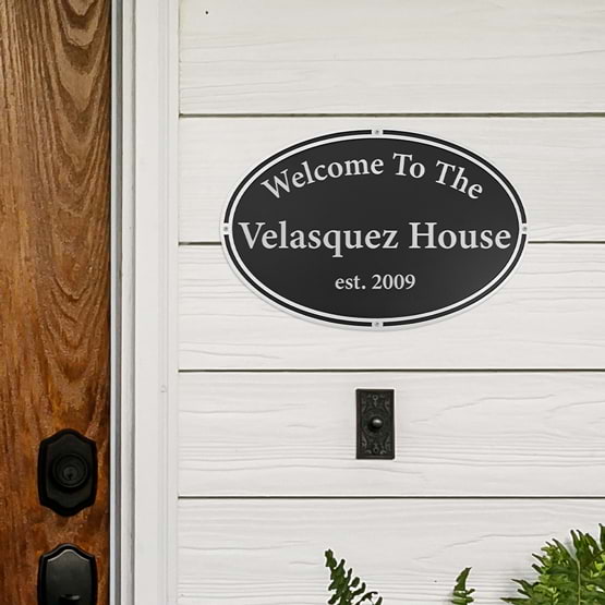 Oval Cast Aluminum Welcome Sign Mounted On Front Porch With Screws And Featuring Black And Silver Design With Family Name