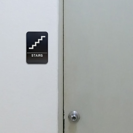 Black And White Plastic Sign With Stairs Icon Above Corresponding Text And Braille Mounted On Wall Beside Stair Access Door