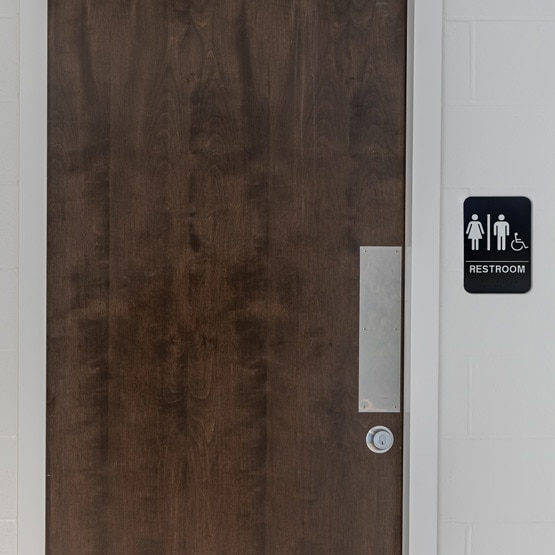 Black And White Plastic Sign With Man And Woman And Wheelchair Icons Hanging On Wall Beside Public Restroom Door