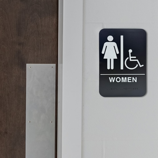 Black And White Plastic Sign With Women And Wheelchair Icons And Text And Braille Mounted On Wall Beside Public Wood Door