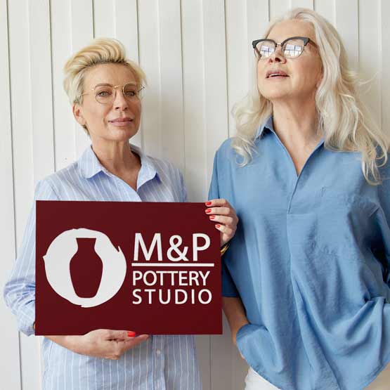 Two Women Business Owners Holding A Maroon Plastic Sign For Their Pottery Studio.